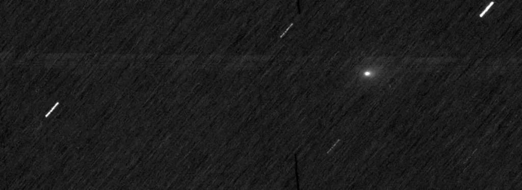 Fond noir de l'espace avec des traînées blanches et un seul point blanc flou. Les stries sont des étoiles et le point est la comète 3I/ATLAS.